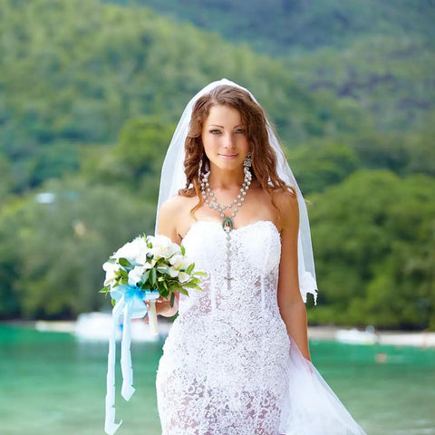 Bride wearing a white lace dress and a rosary necklace, holding a bouquet, standing outdoors with greenery in the background.