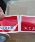 Tamper-evident red security tape roll with white border; serial number label N00006983 visible, held by hand