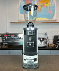 Automatic Espresso Tamper Provides Hella Level Compaction With Adjustable Tamping Pressures Designed To Go Hard On Consistency For Mahlkonig E65 Grinders White 53mm showcased in cafe setting with coffee grinder.