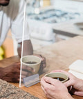 Six Pack Heavy Duty Self Adhesive Sneeze Guard Holders With Secure Locking Design For Quarter Inch Clear Acrylic Sheets in use between two people holding coffee.