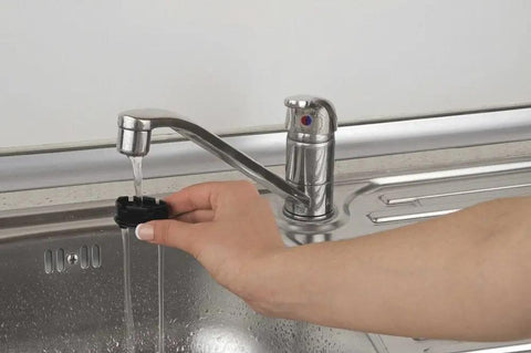 Hand using a faucet sprayer attachment with running water over a stainless steel sink.