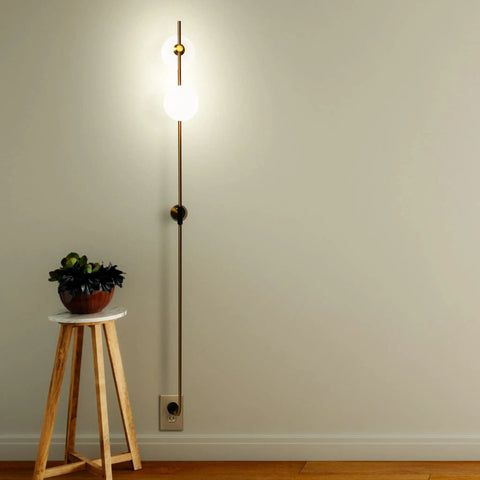 Equinox LED Wall Sconce Indoor Plug In Light For Bedroom And Bathroom With Glass Globe Shades Industrial Wall Mounted Fixture And On Off Switch Gold Finish Perfect For A Modern Home Vibe, shown illuminated on a wall with a side table.