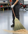 42 Inch Natural Fiber Wire Banded Warehouse Broom With Real Wood Handle For Sweeping Concrete Driveways Garages And Kitchens Built Tough To Handle The Daily Grind being used to sweep wooden debris on a concrete floor.