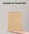 Person holding a small brown kraft paper gift bag with handles, suitable for small gifts, part of 100 Pack Small Brown Kraft Paper Gift Bags With Handles 5.8x3.2x8.25 Inches Natural Kraft Bags For Party Favors Takeout Retail Grocery Weddings Goody Bags And DIY Gifts.