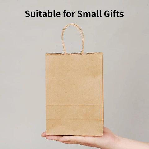 Person holding a small brown kraft paper gift bag with handles, suitable for small gifts, part of 100 Pack Small Brown Kraft Paper Gift Bags With Handles 5.8x3.2x8.25 Inches Natural Kraft Bags For Party Favors Takeout Retail Grocery Weddings Goody Bags And DIY Gifts.