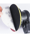 Hand holding a hook and loop sanding disc close to a random orbit sander, showing aluminum oxide paper for smooth finishes.