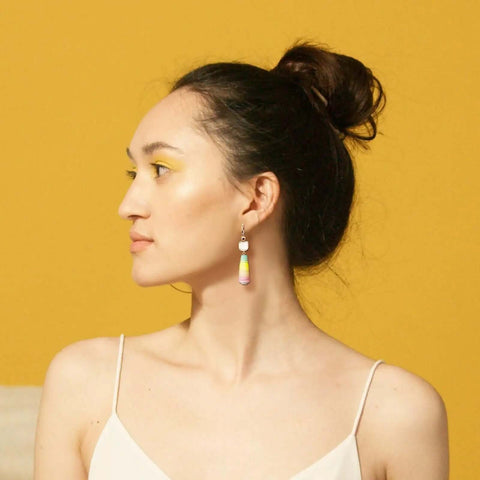 Side profile of a person with updo hairstyle wearing colorful dangle earrings against a yellow background.