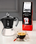 Heavy Duty Moka Induction Coffee Maker With Rock Solid Stainless Steel Boiler And Premium Aluminum Lid Brews Smooth Two Cup Espresso That Nails Your Morning Kickstart In Deep Black with coffee cup and Bialetti coffee bag.