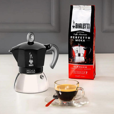 Heavy Duty Moka Induction Coffee Maker With Rock Solid Stainless Steel Boiler And Premium Aluminum Lid Brews Smooth Two Cup Espresso That Nails Your Morning Kickstart In Deep Black with coffee cup and Bialetti coffee bag.