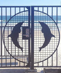 Beach gate with dolphin design leading to sandy beach and ocean view with clear sky.