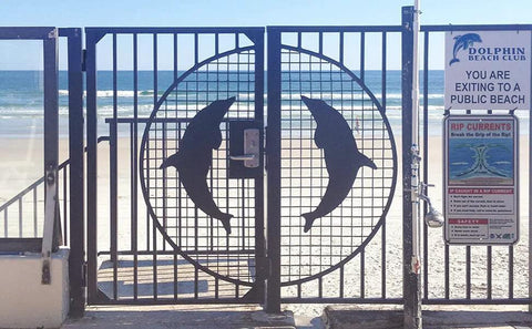 Beach gate with dolphin design leading to sandy beach and ocean view with clear sky.