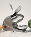 Electric Ice Shaver With Prevent Splash Design 3 Blade Snow Cone Machine 380W Shaved Ice Maker Cranks Out Up To 286 Lbs Per Hour Home Use Ice Crusher With Free Ice Pick Silver displayed on kitchen countertop with fruit and ice cubes.