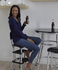 Woman sitting on retro style counter height chair with pull out step stool cushioned vinyl seat and back in black and chrome finish.