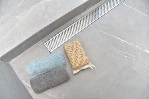 Built Like A Tank Extra Thick Six Gauge Stainless Steel Linear Shower Drain Kit Featuring Brushed Wedge Grate For Tile Floors And Zero Clog Hair Catcher Strainer Fifty Nine Inches Long with towels on tiled floor.