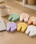 Six Pack Soft Foam Paw Shaped Door Stoppers For Finger Pinch Protection In Pastel Colors Sage Cream Peach Lavender Butter Yellow White on table.