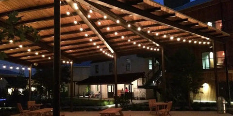 Outdoor patio at night with string lights under wooden canopy, tables and chairs visible, residential buildings in background.