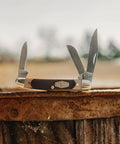 Tough As Nails Three Blade Folding Pocket Knife With High Carbon Stainless Steel Edge And Ergonomic Sawcut Grip displayed on rustic wood.