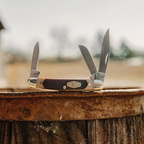 Tough As Nails Three Blade Folding Pocket Knife With High Carbon Stainless Steel Edge And Ergonomic Sawcut Grip displayed on rustic wood.