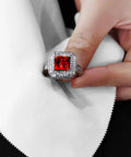 Silver ring with a red square-cut center gemstone surrounded by a halo of small diamonds, held between fingers.