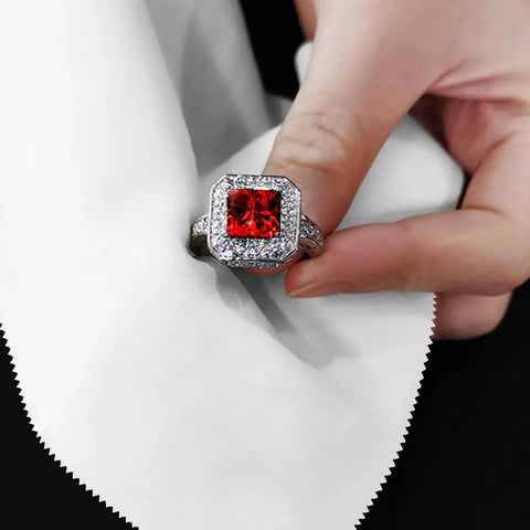 Silver ring with a red square-cut center gemstone surrounded by a halo of small diamonds, held between fingers.