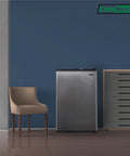 A Top Shelf 3 Cubic Foot Midi Upright Freezer In Stainless Steel With Reversible Door And Adjustable Feet For Basements Or Break Rooms This Sweet Space Saver Delivers Zero Fuss Setup And Drops Heavy Chill Wherever You Park It Today next to a chair.