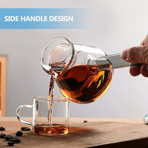 Glass coffee pot with side handle pouring coffee into glass cup, wooden surface, beans scattered