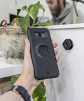 Hand holding phone case near Adhesive Phone Wall Mount Twin Pack For Non Porous Surfaces With 3M VHB Backing Sticks To Any Case Or Universal Adapters No Fuss Easy Permanent Wall Mount Or Screw In Option For A Clean Setup on wall.