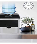 Top Tier Self Cleaning Countertop Ice Maker Portable Electric Machine with water jug on top and plant beside it.