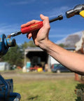 Side view: Grease Buster red-handle tool by LockNLube flushing dried grease from a rusted blue fitting; hammer in hand