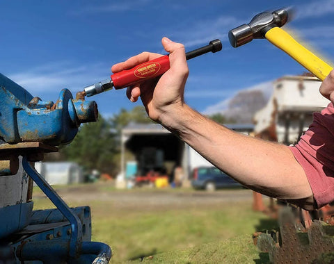Side view: Grease Buster red-handle tool by LockNLube flushing dried grease from a rusted blue fitting; hammer in hand
