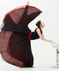 Person in a black and red flowing dress performing a high dance pose with a stool on a white background.