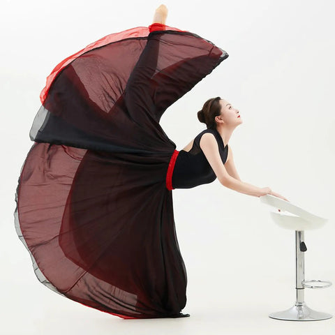 Person in a black and red flowing dress performing a high dance pose with a stool on a white background.