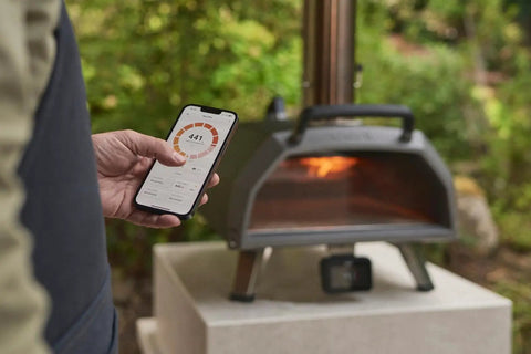 Person checking phone near Karu 2 Pro Multi Fuel Pizza Oven For Backyard Grilling Wood Or Gas Fired 16 Inch Stone Pizza Maker