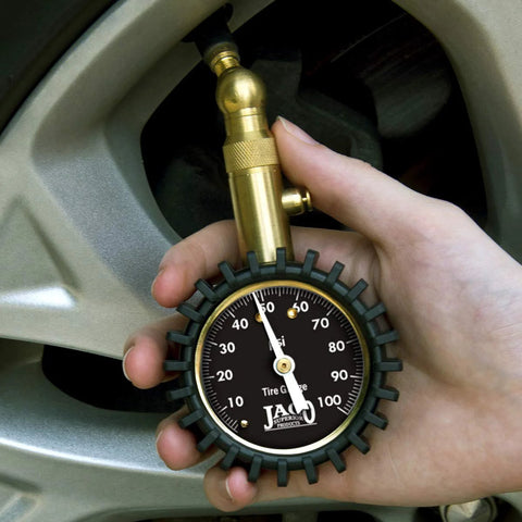 Close-up of brass JACO Elite tire pressure gauge with a 0–100 psi dial held near a tire valve.