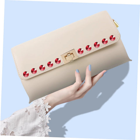 Front view: beige clutch purse with red circular rivet studs along the top edge and a gold clasp, held by a hand.
