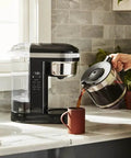 Person pouring from 12 Cup Drip Coffee Maker With Spiral Showerhead And Programmable Keep Warm Plate For Rich Consistent Brews Every Time Onyx Black KCM1209 No Fuss Programmable Timer Auto Shutoff Large Water Reservoir For Busy Mornings