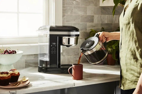 Person pouring from 12 Cup Drip Coffee Maker With Spiral Showerhead And Programmable Keep Warm Plate For Rich Consistent Brews Every Time Onyx Black KCM1209 No Fuss Programmable Timer Auto Shutoff Large Water Reservoir For Busy Mornings