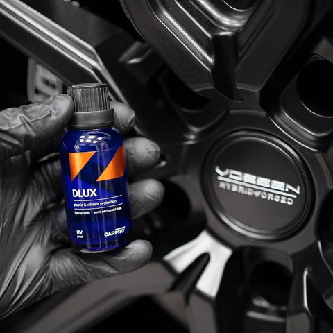 CARPRO DLUX wheel and trim coat bottle (blue) held by gloved hand beside a car wheel