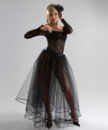 Woman in elegant black tulle gown with lace top and long sleeves, accessorized with a hairpiece, standing against gray background.