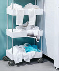 Three-tier laundry cart with white bins filled with clothes against a teal wall near a door.