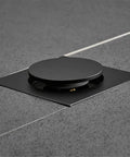 Drop Down Square Pop Up Floor Drain With Matte Black Chrome Cap and Odor Locking Grille For Bathrooms on textured tile floor.