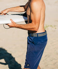 Man wearing Original Unisex Adventure Belt with heavy duty elastic webbing and non metal travel buckle, holding a surfboard.