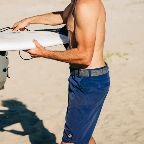Man wearing Original Unisex Adventure Belt with heavy duty elastic webbing and non metal travel buckle, holding a surfboard.
