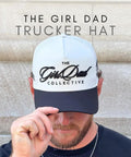 Man wearing The Girl Dad Trucker Hat with white front panel and black bill, holding the brim, against stone background.