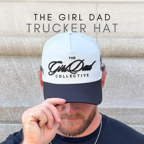 Man wearing The Girl Dad Trucker Hat with white front panel and black bill, holding the brim, against stone background.