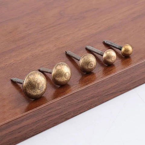 Close-up of five 20-Piece Pack Antique Brass Upholstery Tacks on wood, showing different sizes and threaded steel bodies.