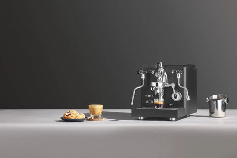 Alto Espresso Machine With Flow Control, Dual Boiler And PID in black next to a cup of espresso and pastries on a tabletop.