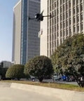 Dual Payload Drop Kit With Emergency Lighting For M30 Drone flying near skyscrapers, visible in clear urban sky with trees.