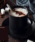 Black cup of mocaccino on dark saucer, topped with whipped cream and cocoa powder, spoon inserted, sugar cubes in background.
