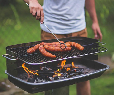 Person grilling sausages on a charcoal grill outdoors; Emergency Fire Starter Pouches For Camping And Outdoor Adventures, Eco Friendly.